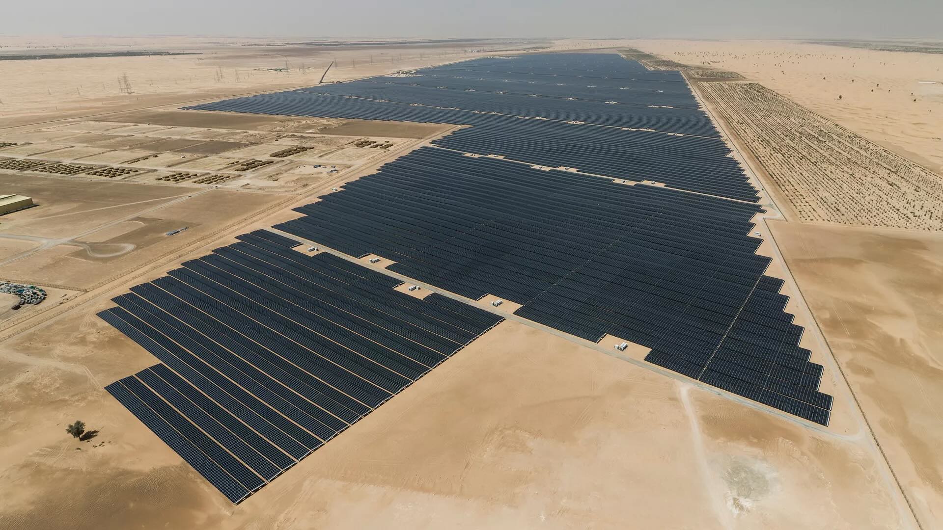A large-scale solar power plant with rows of photovoltaic panels in a desert environment shown from the sky.